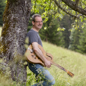 Mann im grauen T-Shirt lehn am Baum und hält seine Gitarre unterm Arm